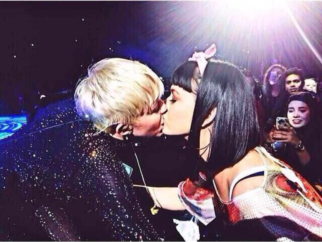 Miley Cyrus bends down to give Katy Perry a kiss